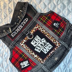 May include: A black denim vest with patches. The vest features red plaid, leopard print, and patches with the text "EXPLOITED", "SUB HUMANZ", and "GBH". Silver studs and a skull pin add detail.