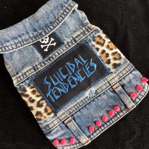 Könnte beinhalten: Eine Jeansweste mit Aufnähern und rosa Nieten. Die Weste hat einen Totenkopf- und gekreuzten Knochen-Aufnäher, einen Leopardenmuster-Aufnäher und einen Aufnäher mit dem Text "SUICIDAL TENDENCIES" in Blau.