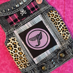May include: A black denim vest with patches, including a pink and black plaid, a leopard print, and a purple logo with the letter "J". The vest has silver studs around the collar and on the bottom.