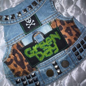 May include: A light blue denim vest featuring silver pyramid studs along the edges, a black patch with a white skull and crossbones, two leopard print fabric panels, and a central black patch with the green embroidered text Green Day.