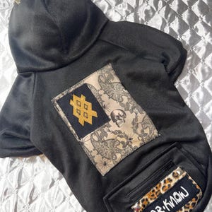 May include: Black dog hoodie with a hood and gold-colored studs. The hoodie features a decorative patch with a skull and a gold-colored design. A pocket with a leopard print patch that reads "DR. KNOW" is also visible.