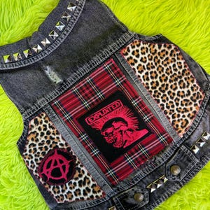 Op de afbeelding: Een zwarte spijkerwester met zilveren studs langs de kraag en zoom. De wester heeft luipaardprint, rode ruit en een rood-zwarte grafische patch met "EXPLOITED". Anarchiesymbool patch.