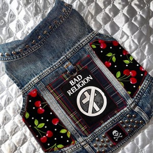 May include: A denim dog vest with silver studs and patches. The vest features a "Bad Religion" patch, a skull and crossbones patch, and cherry-patterned fabric. The vest is designed with a punk rock aesthetic.