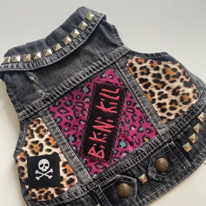 May include: A black denim vest with silver studs, featuring patches of leopard print fabric, pink fabric with the text "Bikini Kill", and a black and white skull and crossbones patch.