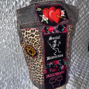 Op de afbeelding: Een grijs spijkergilet met een achterpand voorzien van een hart- en pijlopnaaier, een gedeelte met luipaardprint, een anarchiesymbool-opnaaier en een zwart middenpaneel met bloemaccenten, een skeletafbeelding en de tekst Social Distortion Descendents.