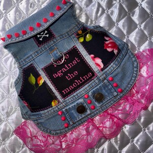 May include: A blue denim vest featuring neon pink cone studs along the collar and hem. The vest includes black floral fabric patches, a metal D-ring, two bronze-colored buttons, and a black patch with the text rage against the machine.