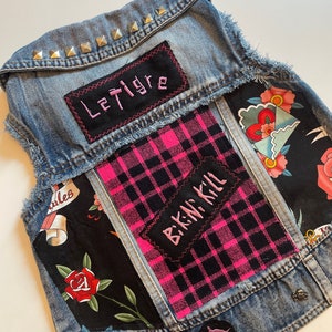 May include: A blue denim vest with patches, including a pink and black plaid patch with the text "Bikini Kill" and a black patch with the text "Le Tigre".