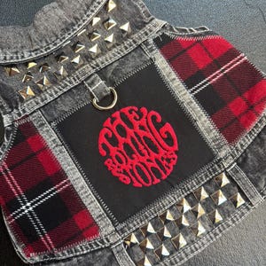 May include: A denim dog vest with a patchwork design featuring red and black plaid fabric. The vest is embellished with silver pyramid studs and a circular patch with the words "Rolling Stones" embroidered in red.