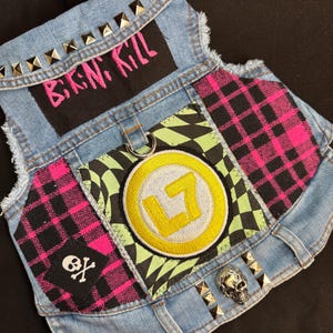 Könnte beinhalten: Eine blaue Hunde-Jeansweste mit Aufnähern und Metallnieten. Die Weste hat einen schwarzen Aufnäher mit dem Text "B.I.K.N. KILL" in Pink, einen Totenkopf-Aufnäher und einen runden Aufnäher mit gelbem "L7"-Logo.