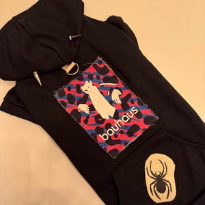 Könnte beinhalten: Schwarzer Kapuzenpullover mit einem bunten Patch mit einer Katze und dem Wort "bauhaus" und einer schwarzen Spinnengrafik auf der Tasche.