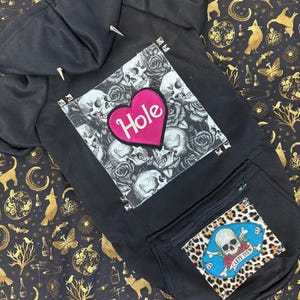 Könnte beinhalten: Schwarzer Kapuzenpullover mit einem Totenkopf- und Rosenmotiv und einem pinkfarbenen Herzen mit dem Wort "Hole" darauf. Der Pullover hat eine Reißverschlusstasche mit einem Totenkopf- und Knochenmotiv und den Worten "Carpe Diem" darauf.