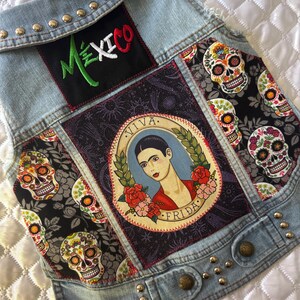 May include: A light blue denim vest featuring silver metal studs along the collar and hem. The back panel displays a black patch with the embroidered text MEXICO, a central oval patch depicting Frida Kahlo with the text VIVA FRIDA, and side panels with a black fabric pattern of colorful sugar skulls.