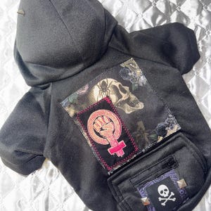 May include: Black dog hoodie with a hood and a pocket. The back features patches with a skull, a feminist symbol, and a skull and crossbones. The hood has metal spikes.