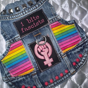 Puede incluir: Un chaleco de mezclilla azul para mascotas con tachuelas piramidales plateadas en el cuello y tachuelas rosas en la parte inferior. Incluye un parche negro con el texto "I bite fascists" en rosa, un parche central negro con un símbolo femenino rosa y rayas arcoíris.