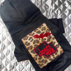 May include: Black dog hoodie with a leopard print patch featuring a red bat and the text "CREATE MODE". The hoodie has a hood with silver spikes.