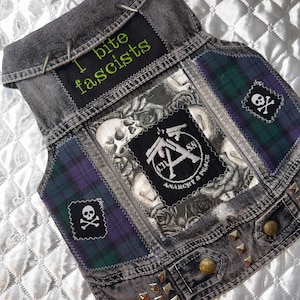 Può includere: Un gilet di jeans grigio con toppe e borchie metalliche. Il gilet presenta una toppa verde e nera con la scritta "I bite fascists". Altre toppe includono teschi, rose e un teschio e ossa incrociate.