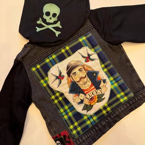 Pode incluir: Uma jaqueta de ganga preta com um remendo xadrez nas costas. O remendo apresenta um marinheiro com uma tatuagem que diz "Lucky". A jaqueta tem um capuz preto com um design de caveira e ossos cruzados verde nas costas.