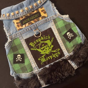 Puede incluir: Un chaleco vaquero para perro con un aspecto desgastado, con un parche de estampado de leopardo con la palabra "ANTIFASCIST". Tiene parches de cuadros verdes, calaveras y tibias cruzadas, y un parche que dice "Dropkick Murphys". El chaleco está ribeteado con piel sintética negra y tachuelas metálicas.