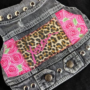 Puede incluir: Un chaleco vaquero gris con tachuelas plateadas en el cuello y los bordes. El chaleco presenta parches de tela de estampado de leopardo con bordados rosas y tela de rosas rosas. Dos botones metálicos aseguran el frente.