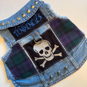 Könnte beinhalten: Eine Hunde-Weste aus Jeansstoff im Punk-Rock-Stil. Die Weste hat einen Totenkopf- und gekreuzten Knochen-Patch, Schottenmuster-Akzente und das Wort "Tendencies" in Blau gestickt. Silberne Nieten verzieren den Kragen und die Kanten, mit einem Metall-D-Ring.