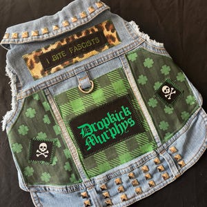 May include: A dog vest made of denim, adorned with patches and metal studs. The vest displays a leopard print patch with the text "I BITE FASCISTS" and a green plaid patch with "Dropkick Murphys". Additional patches include shamrocks and a skull and crossbones.
