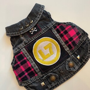 May include: A black denim vest with a yellow circle patch with a white letter L inside. The vest has pink and black plaid fabric patches and silver studs.