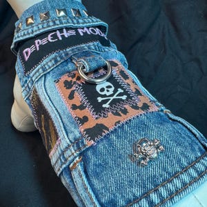 May include: A denim dog vest with a punk rock aesthetic. The vest features a skull and crossbones patch, a metal skull pin, and the text "DEPECHE MODE". The collar is studded with silver metal studs.