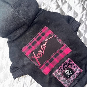May include: Black dog hoodie with a hood and ribbed cuffs. Features a pink and black plaid patch with the text "Xray Apex" and a smaller patch with a skull and crossbones on a leopard print background.
