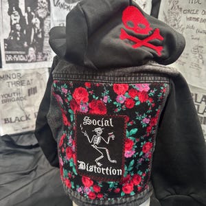 May include: Black hooded denim jacket with a red skull and crossbones on the hood. The back features a floral pattern with a patch that reads "Social Distortion" with a skeleton graphic. The jacket has a punk rock aesthetic.