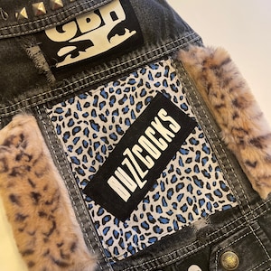 May include: A black denim jacket with patches, including a leopard print fabric, a black patch with the text "Buzzcocks", and a white patch with the text "GBH". The jacket has silver studs on the collar and a skull and crossbones patch on the bottom.