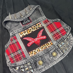 May include: A gray denim vest with metal studs and spikes. The vest features patches of leopard print, red plaid, and a black patch with the text "Cock Sparrer" and "the rich". Two brass-colored buttons are on the front.