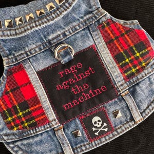 May include: A denim vest with silver stud accents and red plaid patches. The vest features a black patch with the words "rage against the machine" in pink, and a skull and crossbones patch.