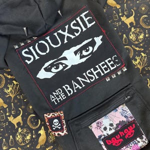 Könnte beinhalten: Schwarzer Kapuzenpullover mit einem großen weißen Patch, auf dem "Siouxsie and the Banshees" steht, einem Totenkopf- und Knochen-Patch auf der linken Seite und einem rosa-schwarzen Patch, auf dem "Bauhaus" steht.