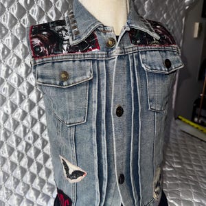 May include: A light blue denim vest featuring a collar with silver studs, two chest pockets with buttons, and a front button closure. The vest is decorated with patches including a bat, a skull, and dark patterned fabric on the shoulders.