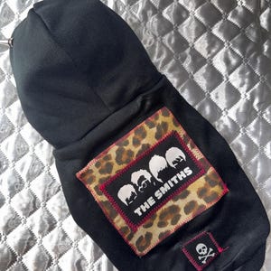 May include: Black dog hoodie with silver spikes on the hood. The back features a leopard print patch with a band silhouette and the text "THE SMITHS". A small skull and crossbones patch is also visible.