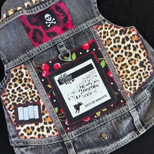 May include: A gray denim vest with gold stud accents and patches. Patches include a skull and crossbones, leopard print, and a black and white graphic with the text "BUG LIFE OUT OF BREATH".