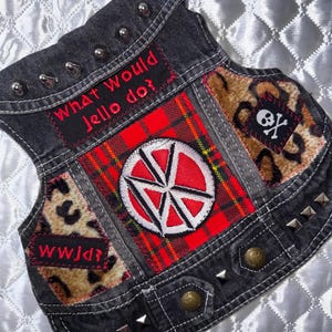 May include: A black denim vest featuring silver metal studs, a red and black plaid patch with a circular white and red logo, leopard print fabric accents, and two black patches with red text reading "What Would Jello do?" and "WWJd?" with a skull and crossbones.