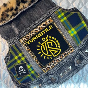 Könnte beinhalten: Eine schwarze Jeansweste mit Leopardenkragen und Aufnähern. Die Weste hat einen großen Aufnäher mit dem Wort "TURNSTILE" und einem gelben Sonnendesign. Zusätzliche Aufnäher sind ein Totenkopf mit gekreuzten Knochen und ein blau-grünes Karomuster. Silberne Nieten verzieren die Weste.