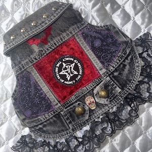 May include: A dark denim vest with a red velvet central panel and purple patterned sections. It has metal studs, decorative buttons, and a black lace trim. The text "THE SISTERS OF MERCY" is on a patch.