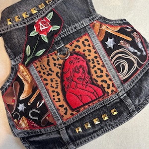 Puede incluir: Un chaleco vaquero negro con tachuelas doradas y varios parches. Los parches incluyen una rosa roja, un estampado de leopardo, una mujer con una camisa roja y un diseño de temática vaquera con botas, una herradura y una cuerda.