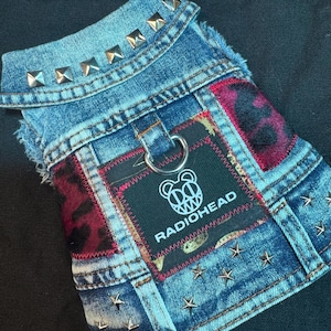 Könnte beinhalten: Eine blaue Hunde-Jeansweste mit silbernen Stern- und quadratischen Nieten. Die Weste hat einen schwarzen Aufnäher mit dem Radiohead-Logo und -Text sowie Flicken aus rot-schwarzem Leopardenmuster-Stoff.