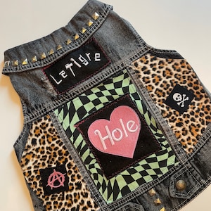 May include: A black denim vest with patches, studs, and a heart-shaped patch that says "Hole". The vest has a leopard print fabric patch, a black and white checkered fabric patch, and a black patch with an anarchy symbol. The vest also has a black patch with the text "Le Tigre" in white.