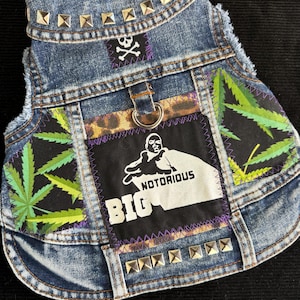 May include: A denim dog vest with silver stud accents and patches. The patches feature a skull and crossbones, cannabis leaves, and a graphic of the Notorious B.I.G. with the word "BIG".