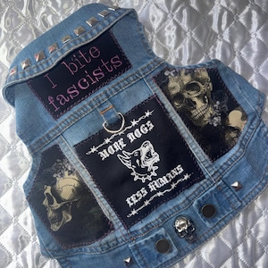 May include: A light blue denim dog vest with patches and silver studs. The vest features patches with skull graphics and the phrases "I bite fascists" and "More Dogs Less Humans".