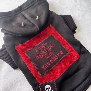 Może przedstawiać: Czarna bluza z kapturem dla zwierząt z czerwoną naszywką z napisem "rage against the machine" w czerwonym haftem. Bluza ma naszywkę z czaszką i skrzyżowanymi piszczelami oraz dwa srebrne kolce na kapturze.