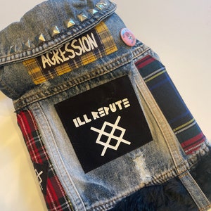May include: A blue denim jacket with patches, including a yellow and black plaid patch, a black patch with the text "ILL REPUTE" and a white symbol, and a pink button with a skull and crossbones. The jacket has silver studs on the collar and a black fur trim.