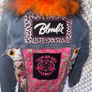 May include: A blue denim jacket featuring a bright orange faux fur collar. The back displays a black patch with the white text Blondie over a pink leopard print, and a central black patch with pink text Cute as Hell over a pink rose pattern.