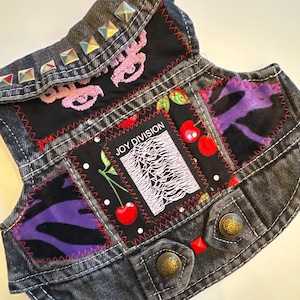 May include: Black denim vest with a variety of patches, including a Joy Division Unknown Pleasures patch, a cherry print patch, and a purple and black animal print patch. The vest has silver studs on the collar.