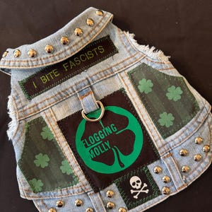 Peut inclure: Un gilet pour chien en jean, orné de clous argentés et de patchs variés. Les patchs présentent un trèfle vert, les mots "FLOGGING MOLLY" et la phrase "I BITE FASCISTS". Un crâne et des os croisés sont également présents.