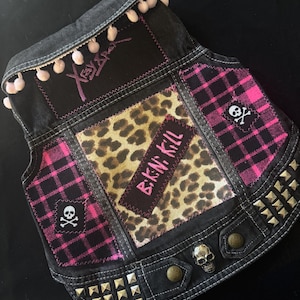 May include: Black denim vest with pink plaid and leopard print patches. Features silver studs, skull accents, and the text "Bikini Kill". The vest is embellished with pink pom-poms and embroidered text on the back.